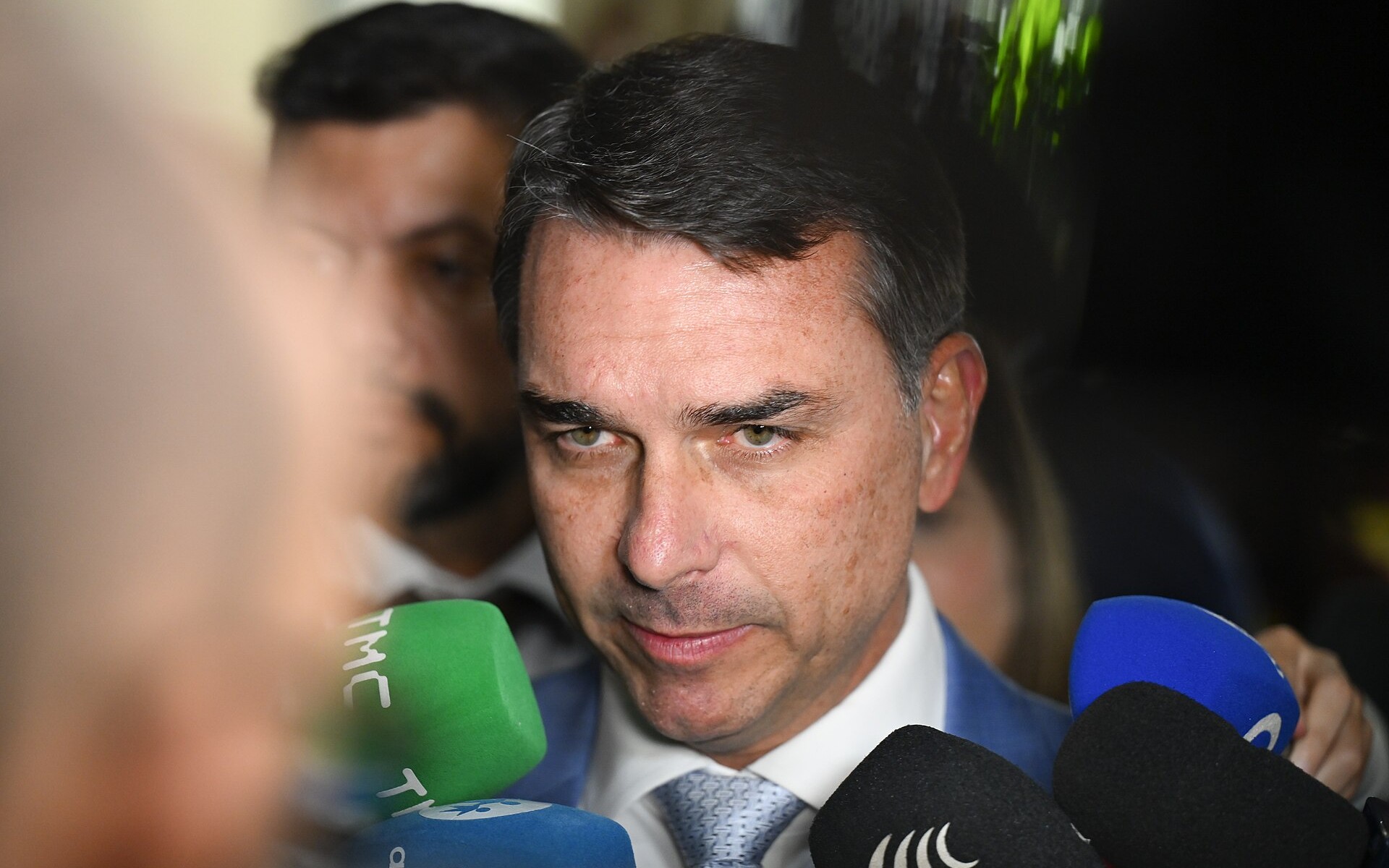 Flávio Bolsonaro, de traje, rodeado de micrófonos