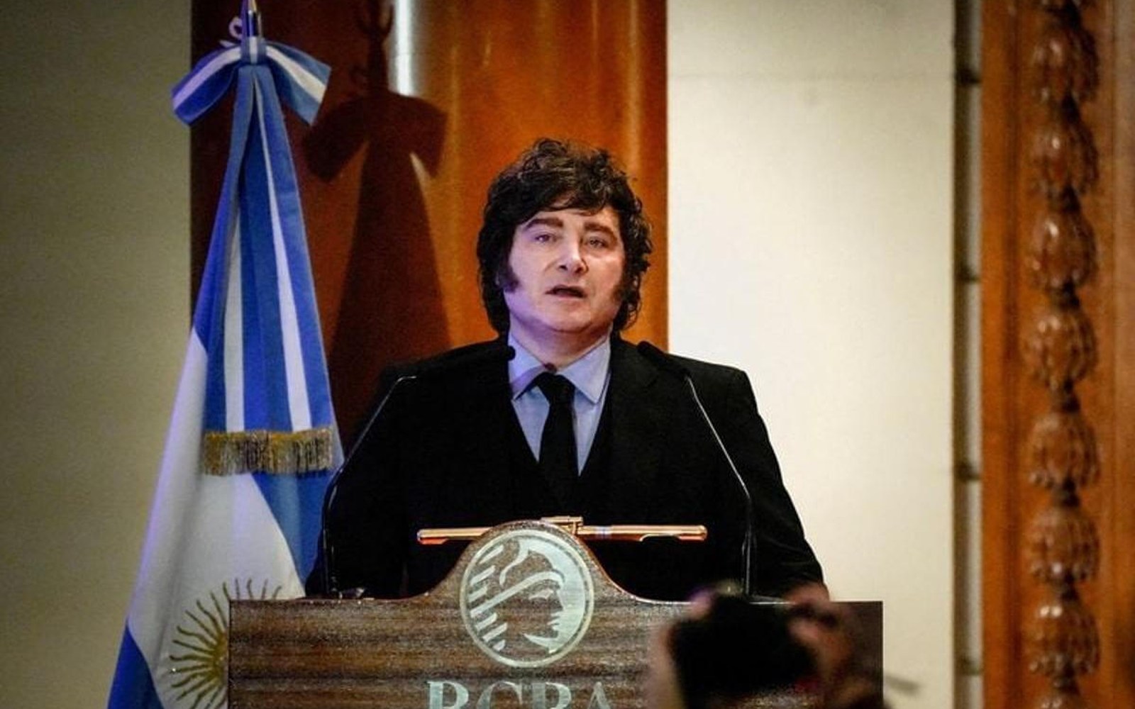 Javier Milei habla en un atril, con la bandera argentina detrás
